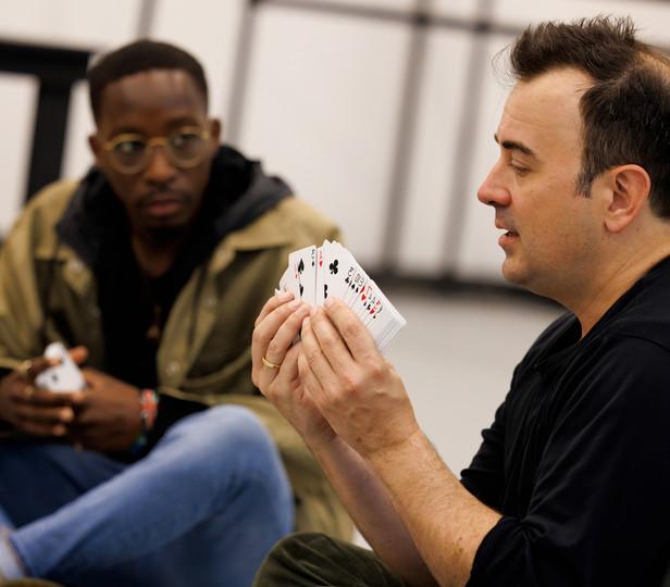 Steve Cuiffo is in a navy shirt holding and demonstrating using a deck of cards. A student, seated on the floor, observes intently. The setting is informal and educational, suggestive of a hands-on learning experience.